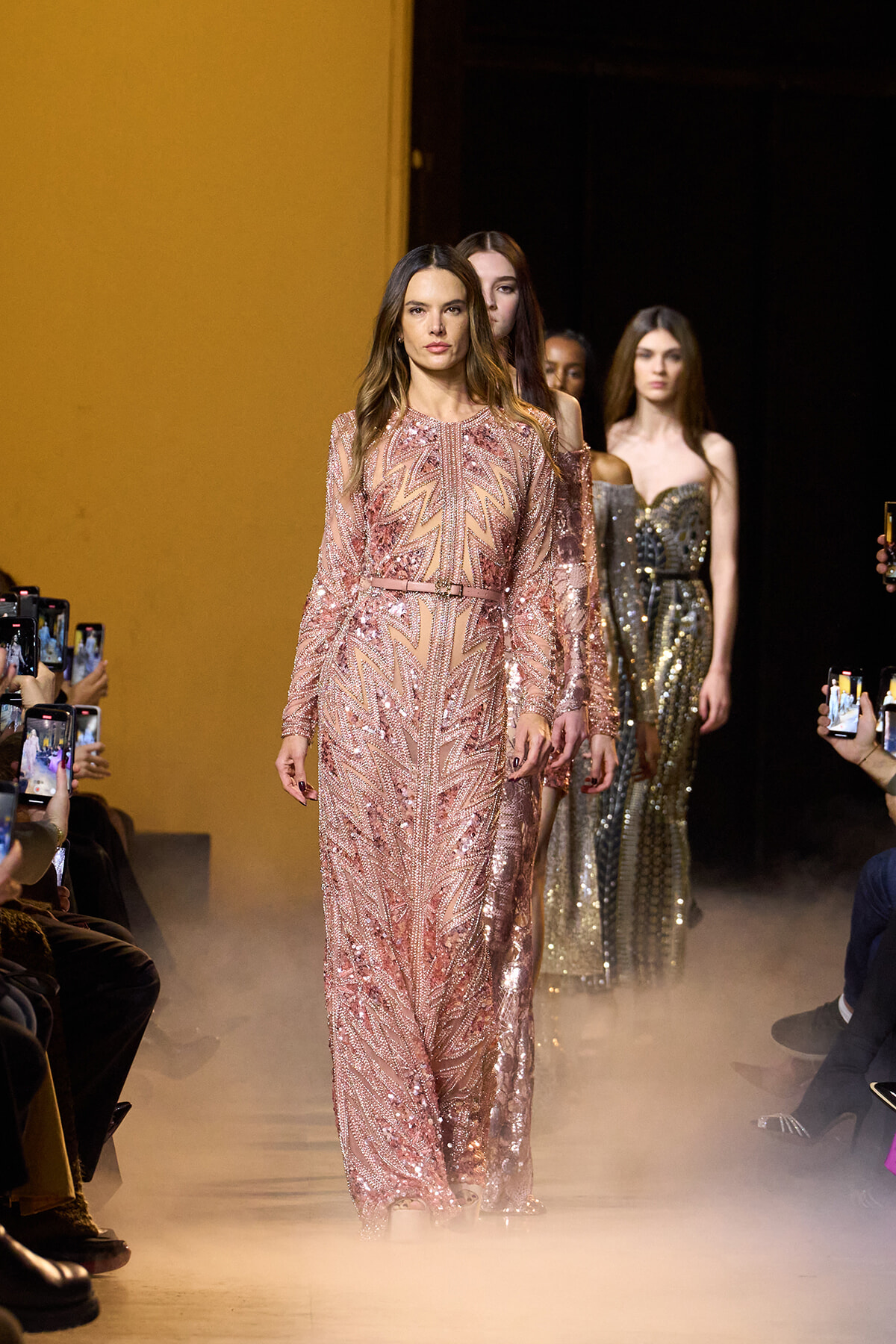 Front-model in a pink sequined gown walks the runway, followed by other models, as audience members take photos nearby.