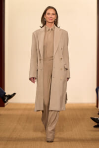 Model on a runway wearing a tonal beige suit: long double-breasted coat over matching shirt and trousers, walking forward.