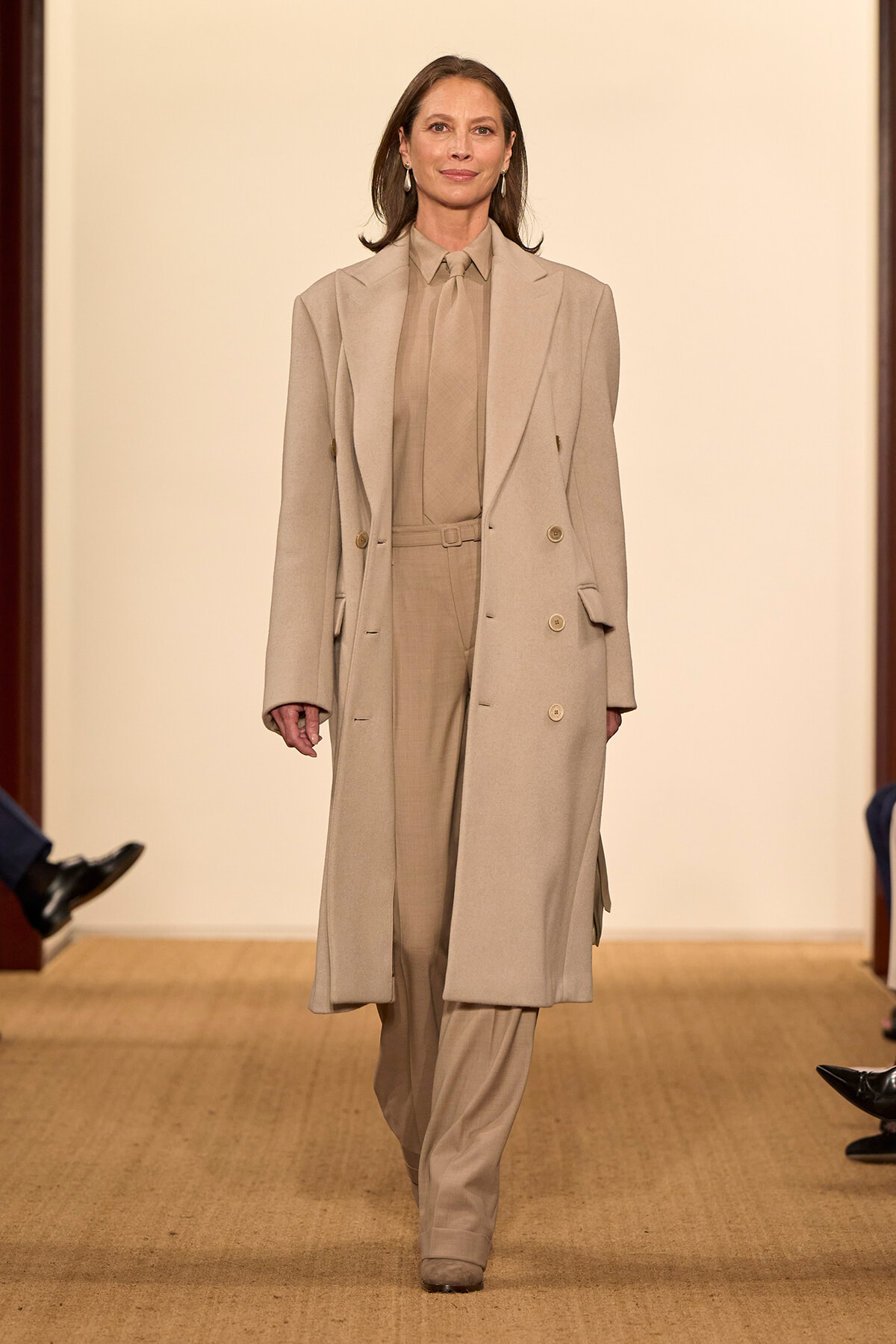 Model on a runway wearing a tonal beige suit: long double-breasted coat over matching shirt and trousers, walking forward.