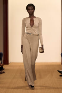 Model walking a beige knit jumpsuit with belt and matching clutch on a runway, neutral-toned ensemble.