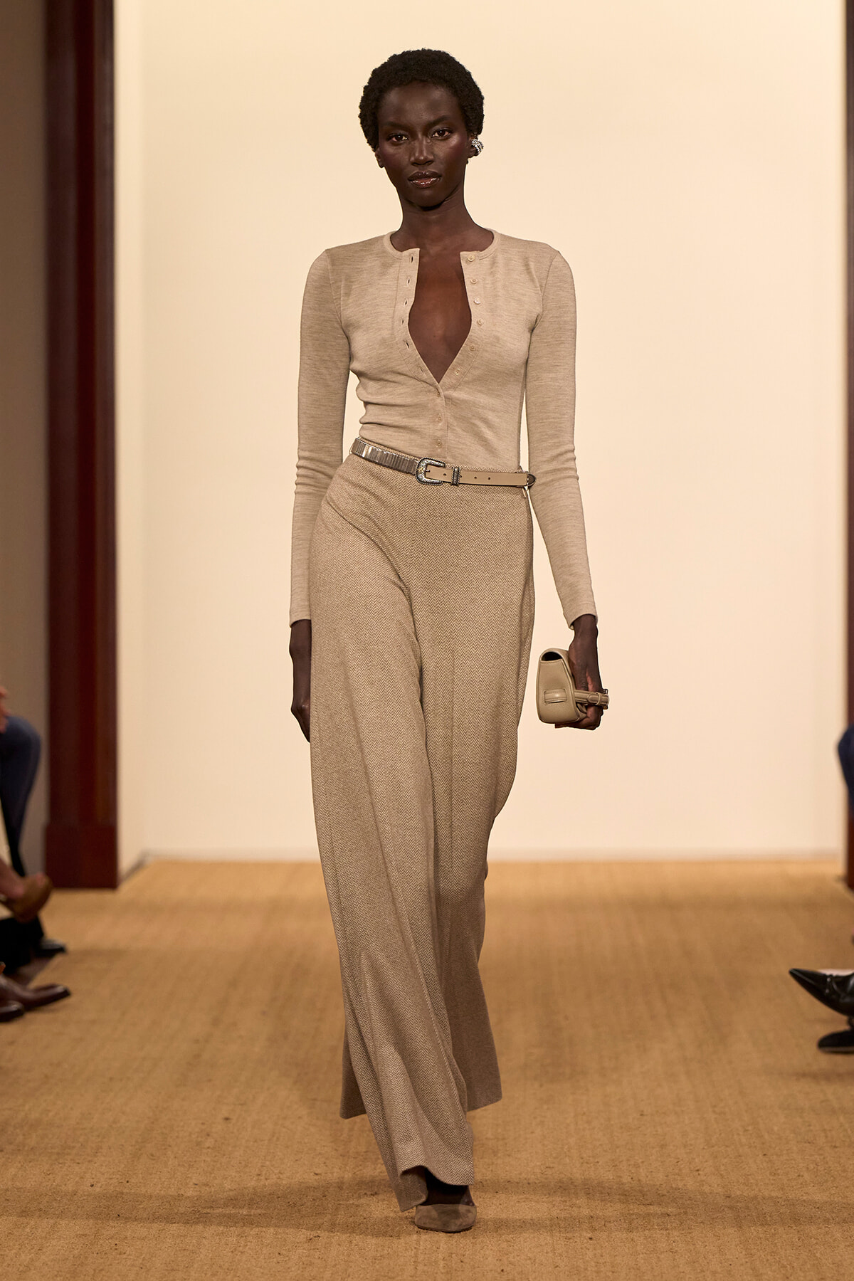Model walking a beige knit jumpsuit with belt and matching clutch on a runway, neutral-toned ensemble.