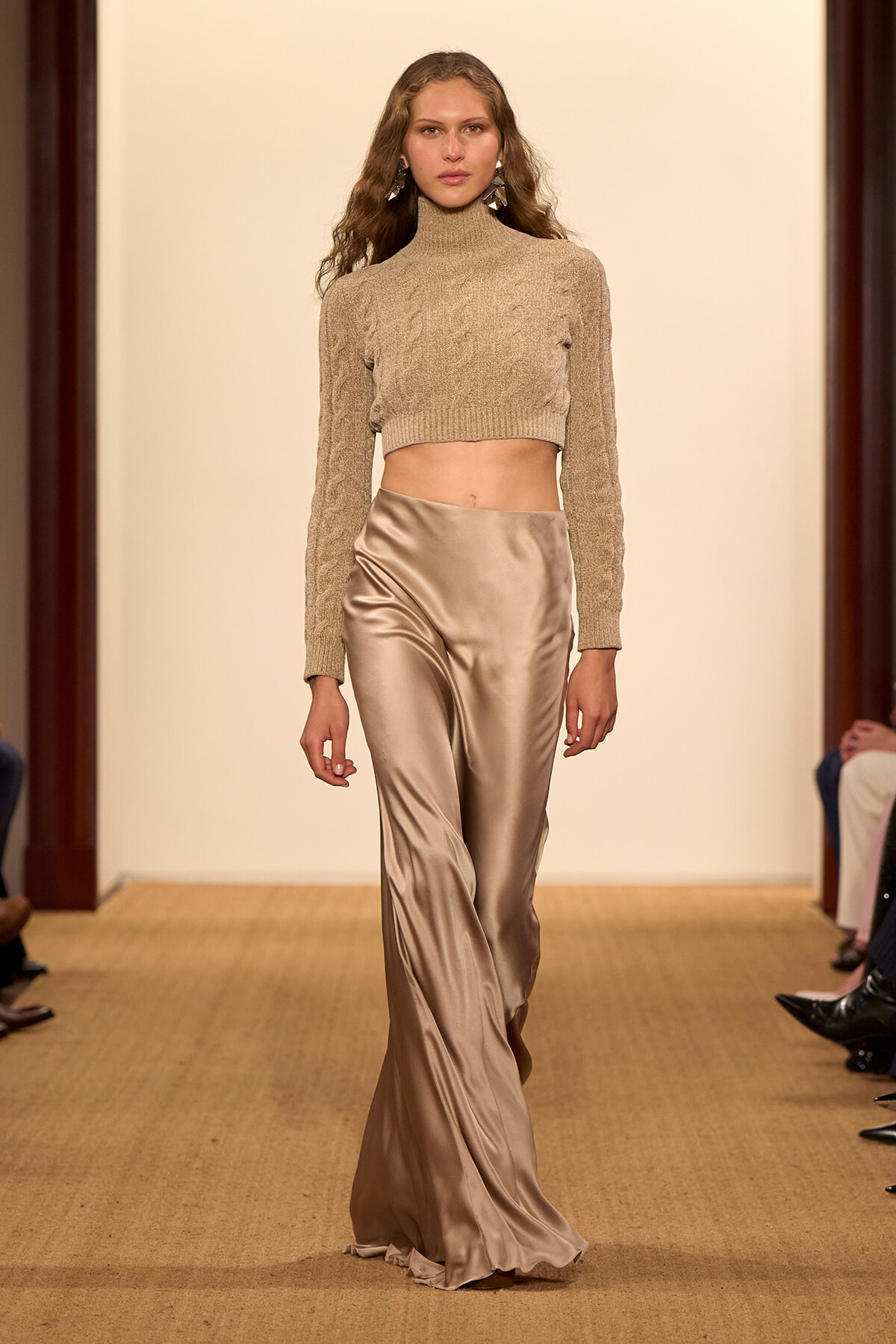 Model walks the runway in a beige cropped knit turtleneck and satin wide-leg trousers.