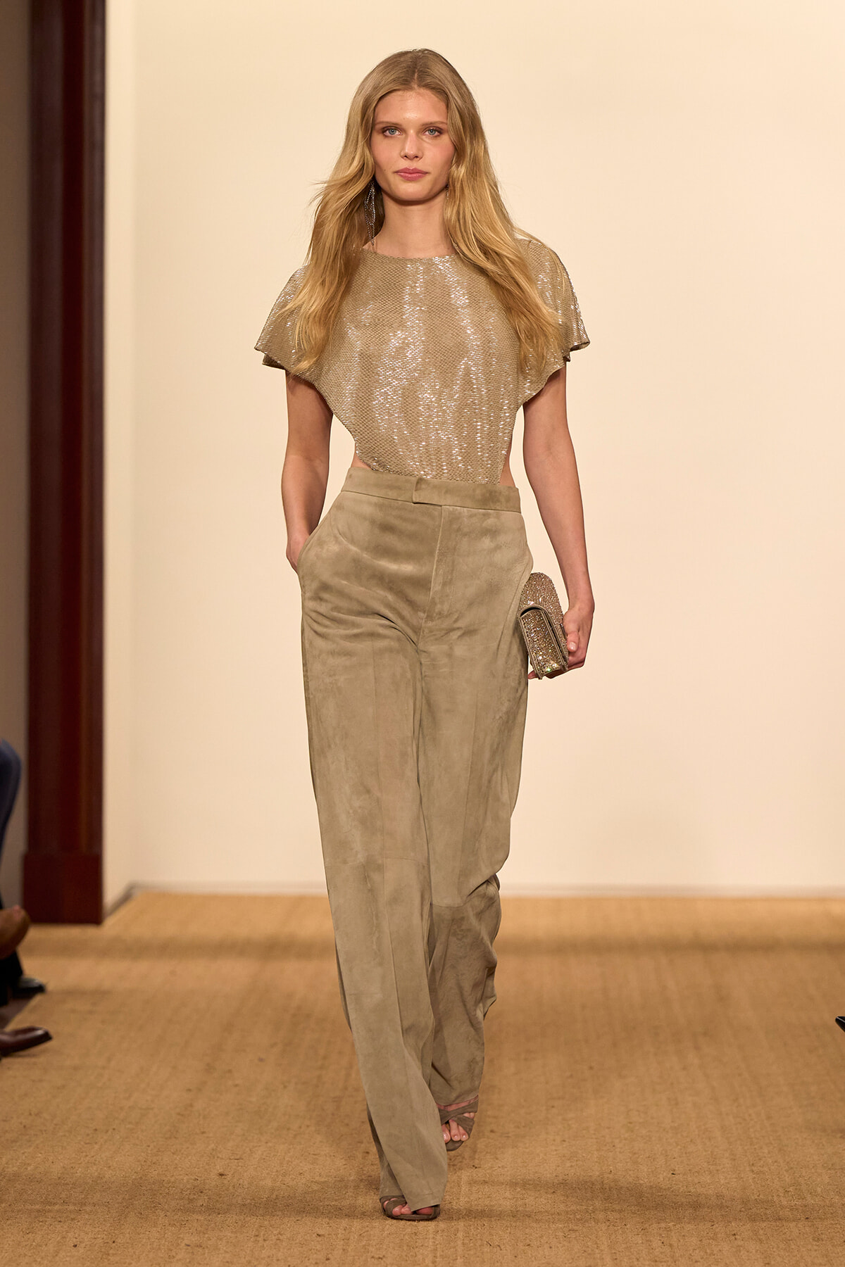 Model walking a runway in a shimmering beige top and light tan suede trousers, carrying a small glittery clutch.