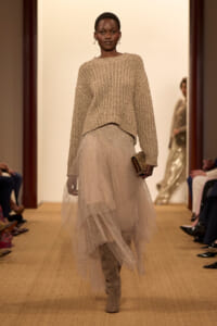 Model walks the runway in a chunky beige knit sweater layered over a sparkly tulle gown, wearing taupe knee-high boots and carrying a small gold clutch.