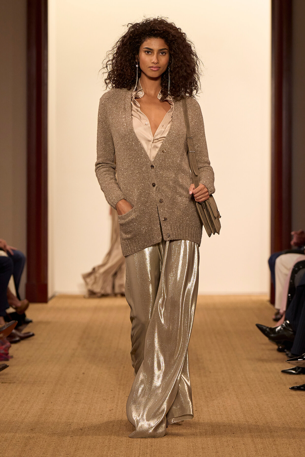Model walking a runway in a beige glitter cardigan over a satin blouse and metallic silver wide-leg pants, carrying a taupe bag.