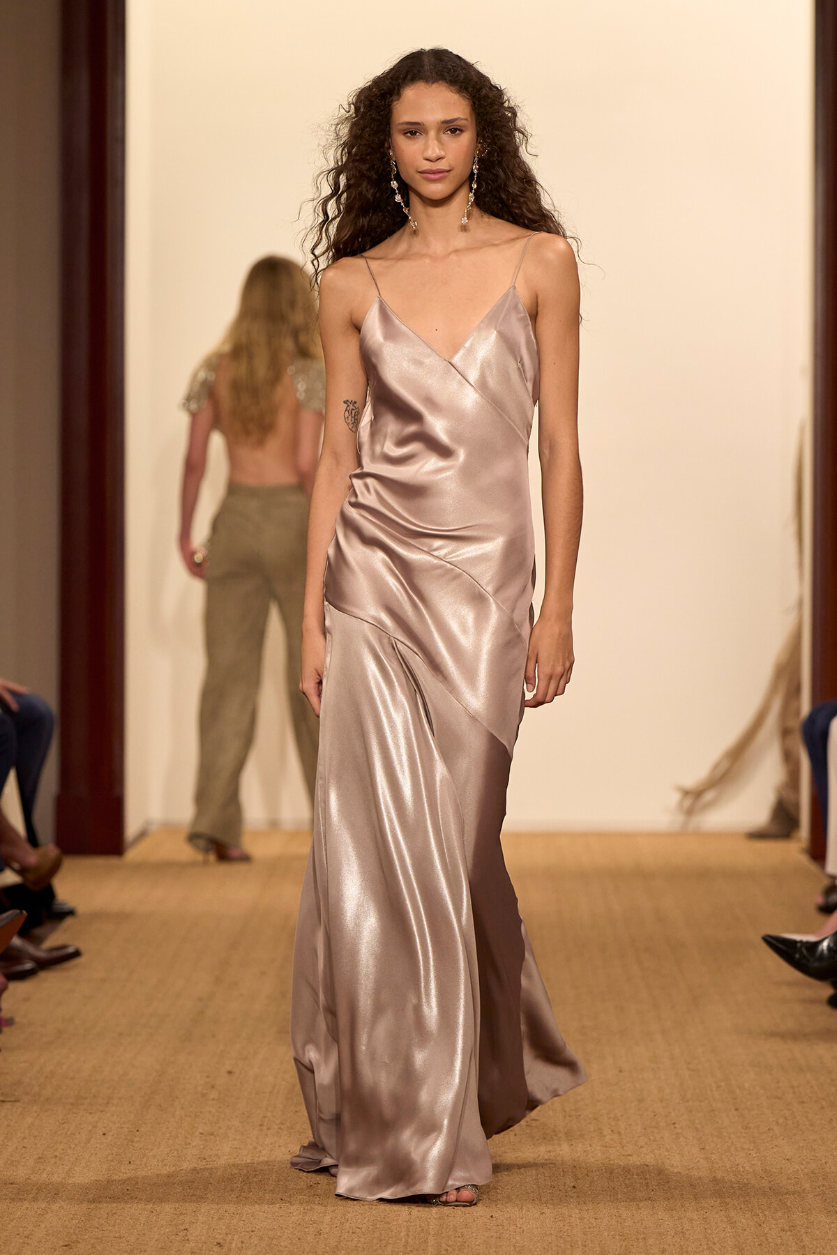 Model walks the runway in a champagne satin slip dress with thin straps and a draped skirt, jewelry visible.
