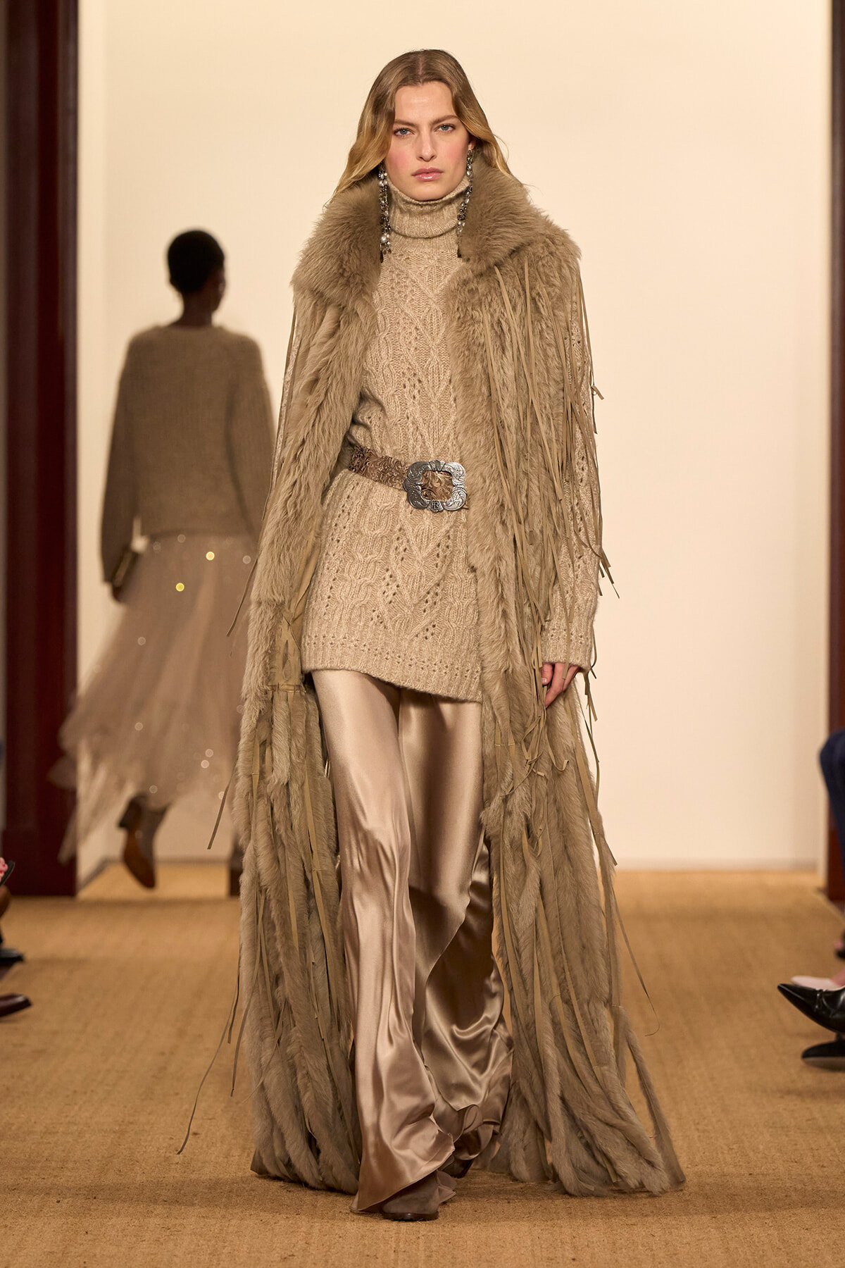 Model walking runway in beige knit turtleneck dress with wide belt and long fringe coat over satin pants.