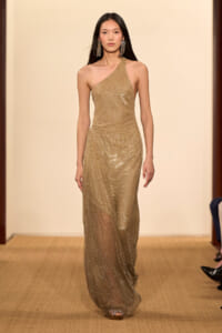 Model walking a runway in a gold one-shoulder sequined gown with statement earrings on a neutral backdrop runway stay.