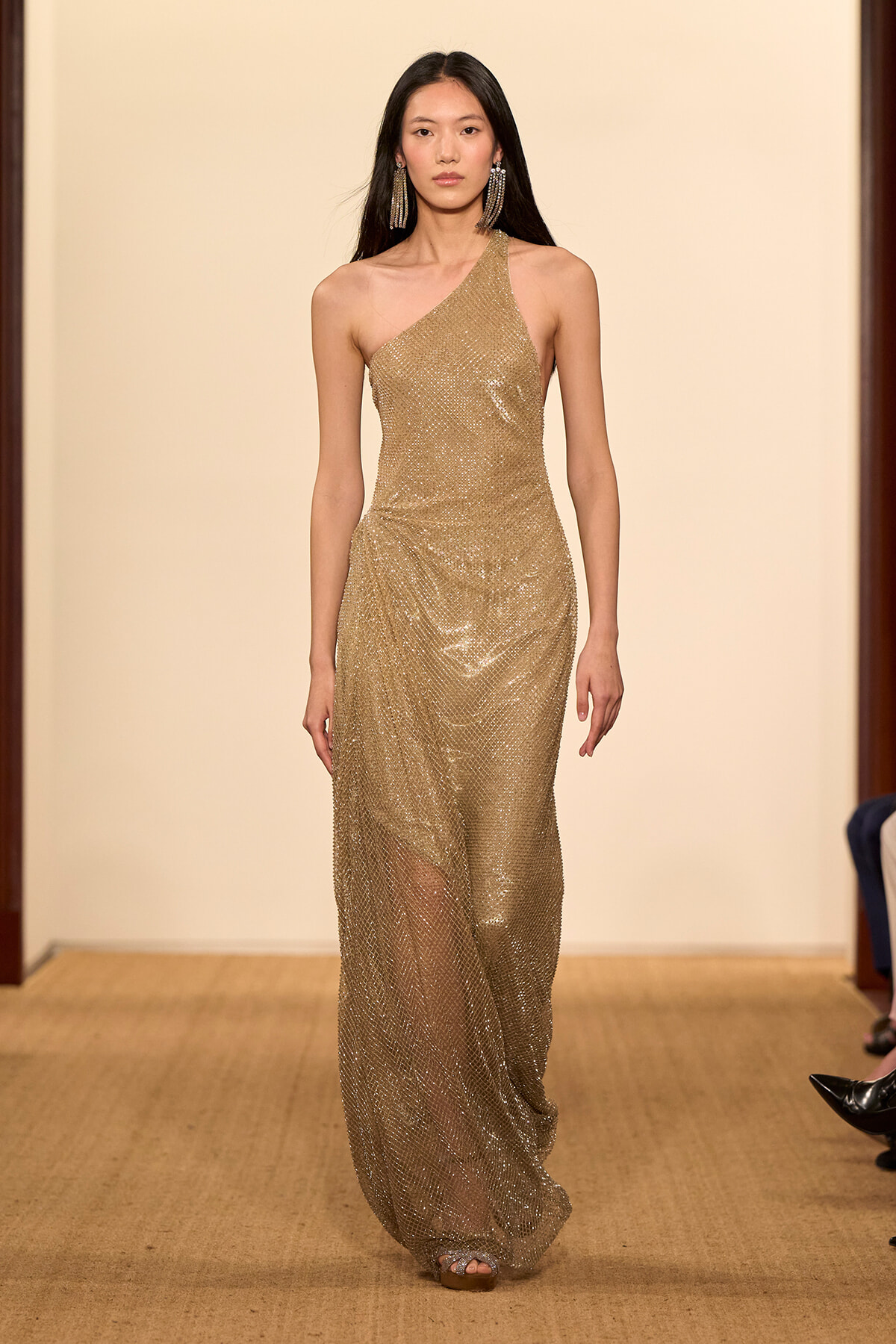Model walking a runway in a gold one-shoulder sequined gown with statement earrings on a neutral backdrop runway stay.