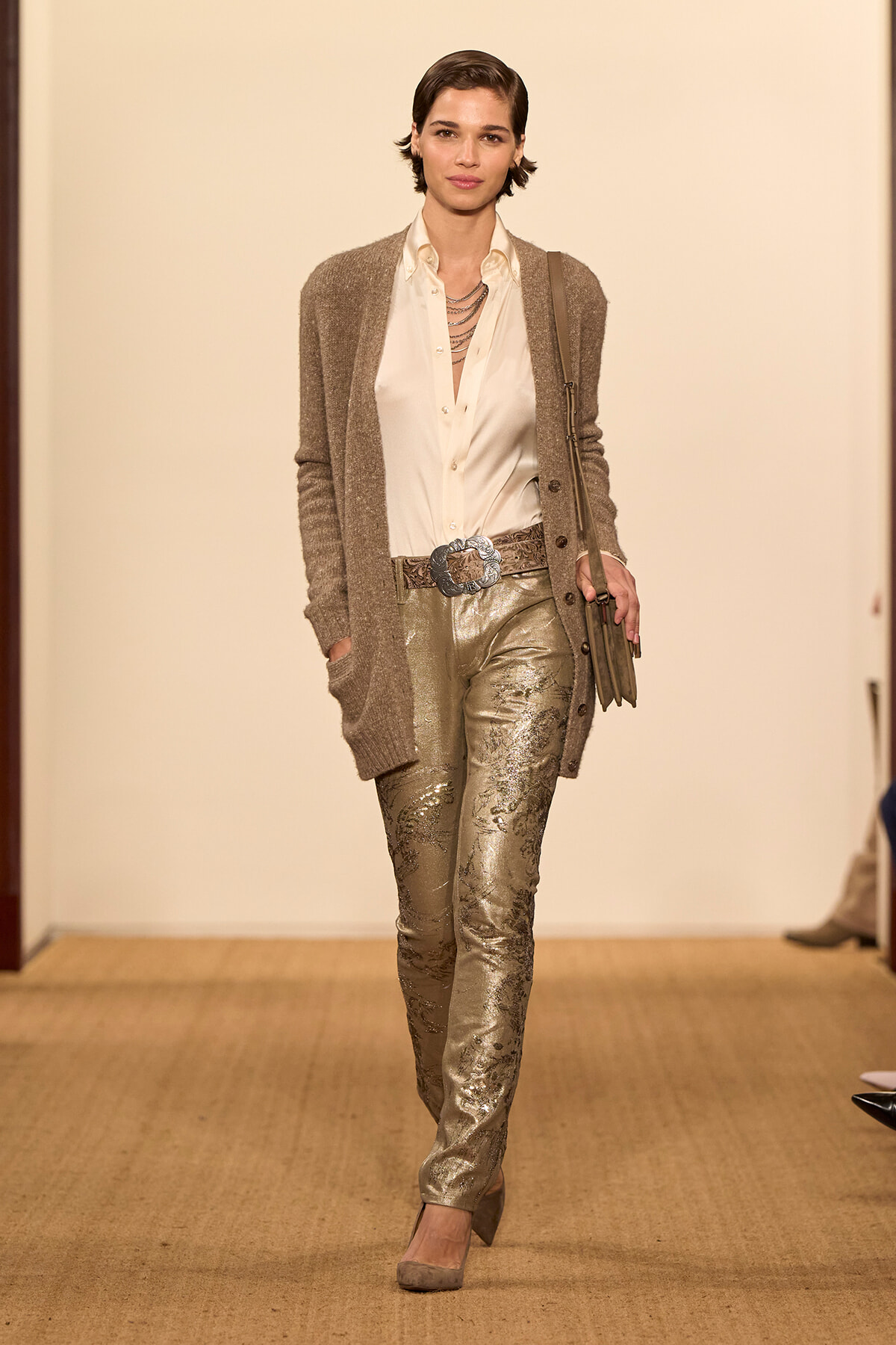 Model on a runway wearing a white blouse, metallic gold pants, and a taupe cardigan with a large belt and layered necklace.