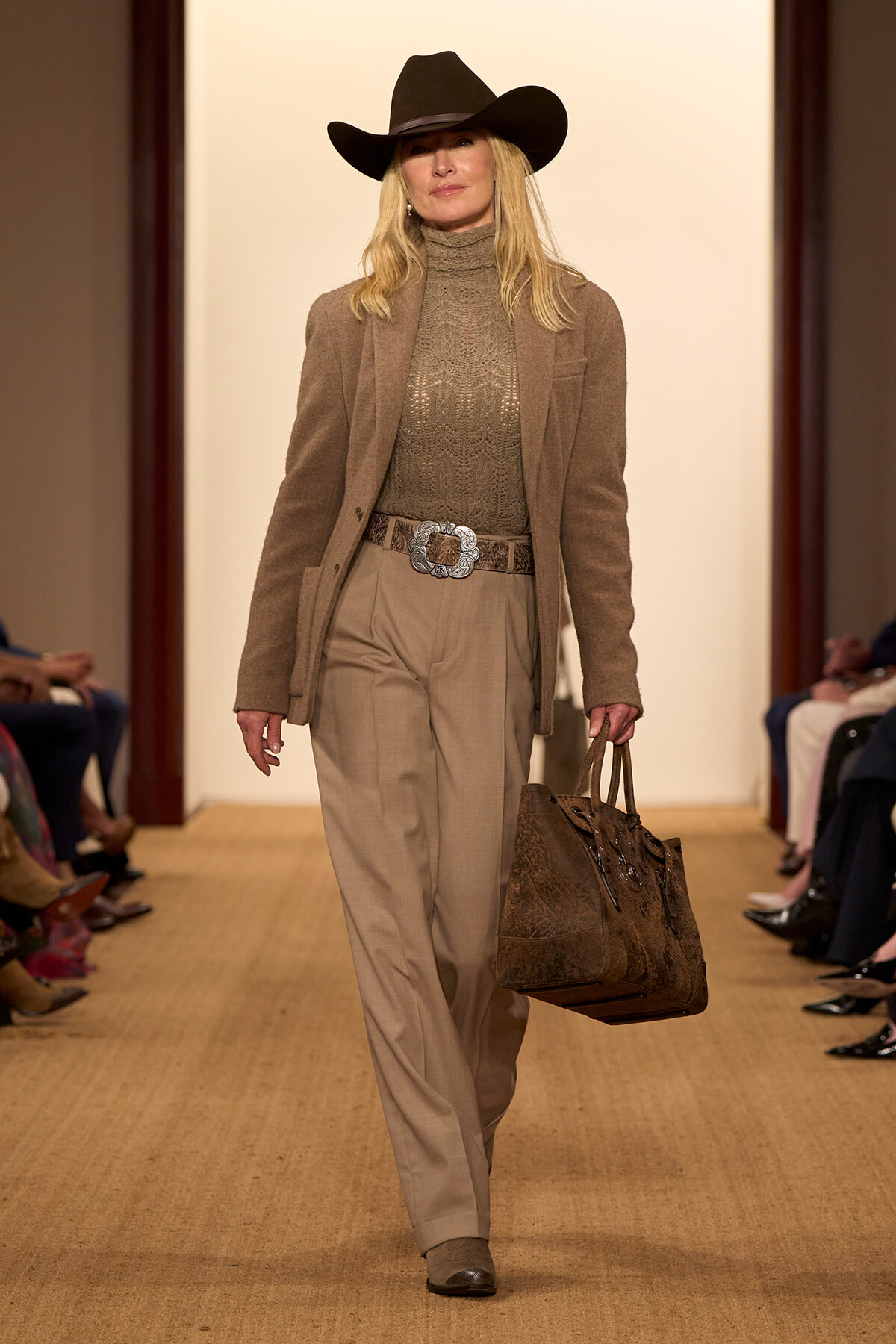 Runway model in a tan Western-inspired outfit: wide-brimmed black cowboy hat, knit turtleneck, blazer, belt, and a large brown handbag.