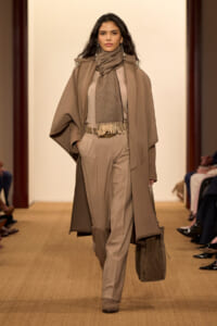 Model walks the runway in a camel coat, matching scarf, and taupe tailored pants in neutral tones.