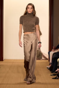 Model walking runway in a brown fuzzy crop turtleneck and champagne satin wide-leg pants, accessorized with long earrings and a fur handbag.