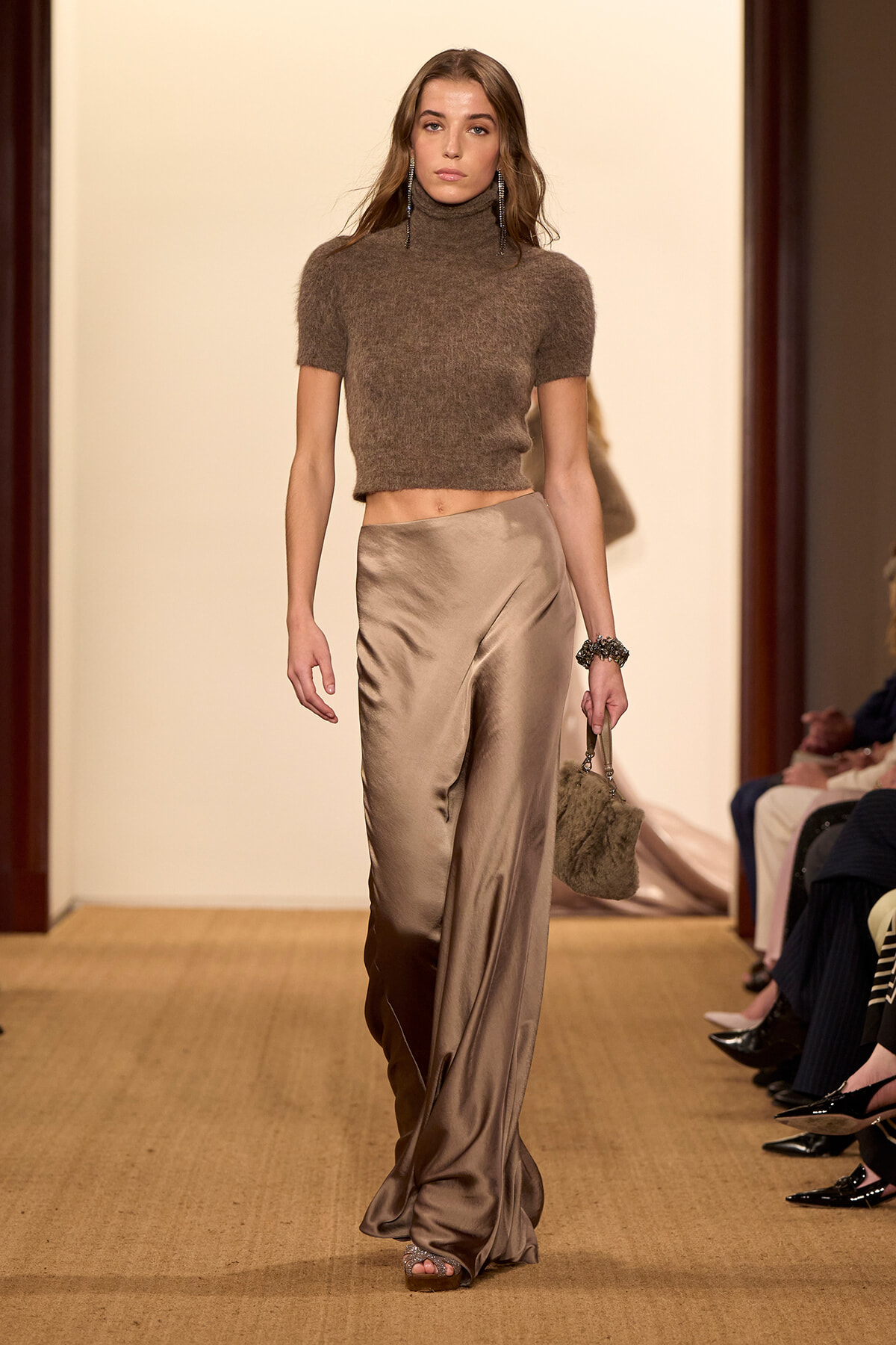 Model walking runway in a brown fuzzy crop turtleneck and champagne satin wide-leg pants, accessorized with long earrings and a fur handbag.