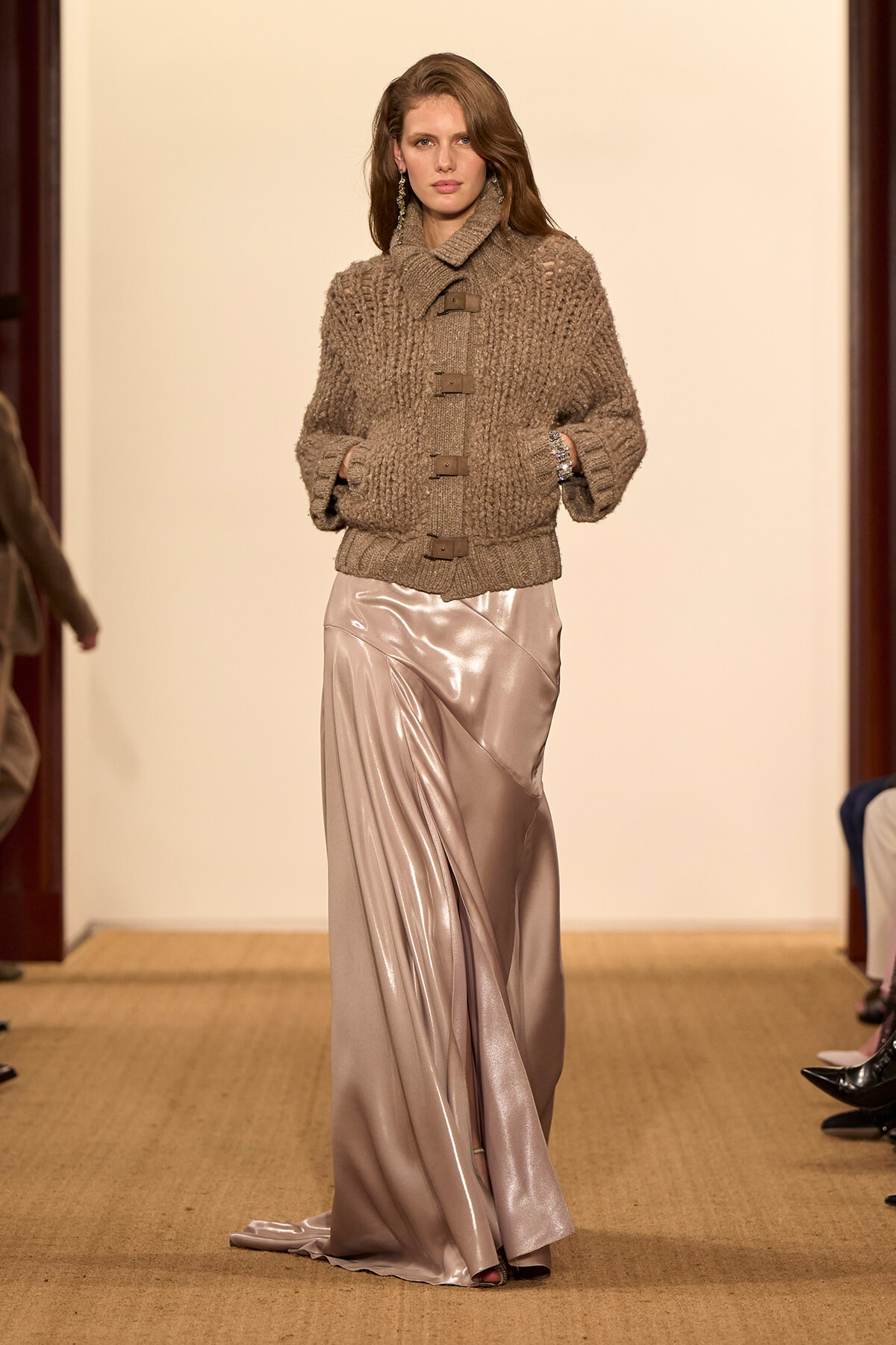 Model walking a runway in a chunky taupe knit cardigan paired with a glossy satin skirt, hands in pockets.