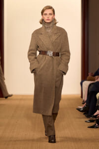 Model walks a runway wearing a brown herringbone/tweed wrap coat cinched with a wide belt featuring an 'FR' buckle, paired with a sequined turtleneck, in a fashion show setting.