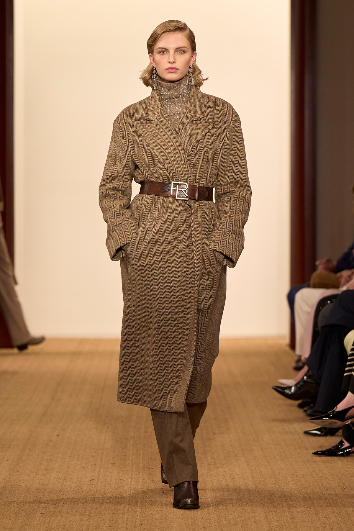 Model walks a runway wearing a brown herringbone/tweed wrap coat cinched with a wide belt featuring an 'FR' buckle, paired with a sequined turtleneck, in a fashion show setting.
