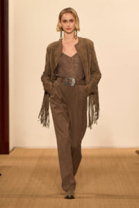 Model walking a runway in brown fringe-sleeve jacket over a sequined brown top and high-waisted trousers with a wide belt and large silver buckle.
