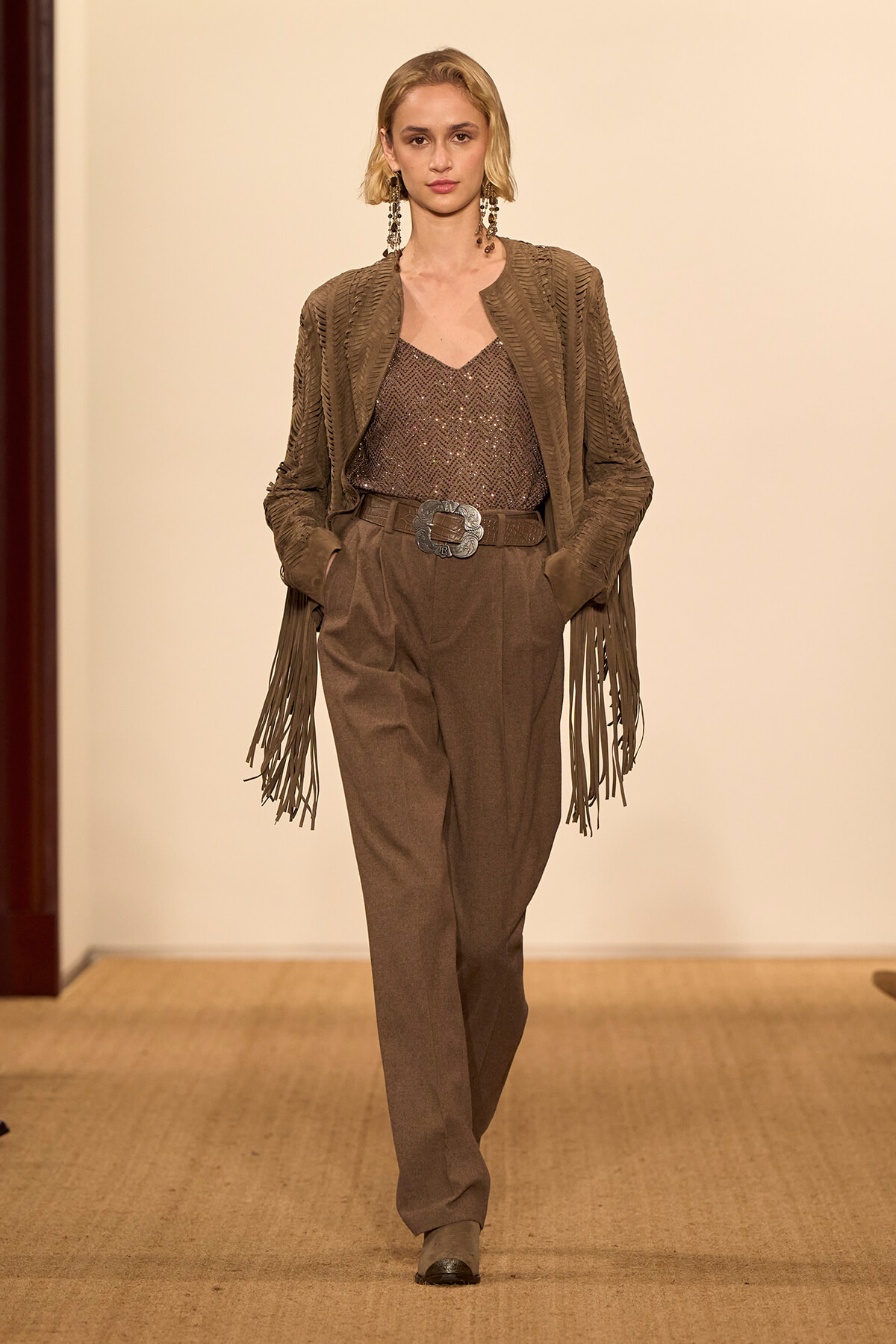 Model walking a runway in brown fringe-sleeve jacket over a sequined brown top and high-waisted trousers with a wide belt and large silver buckle.