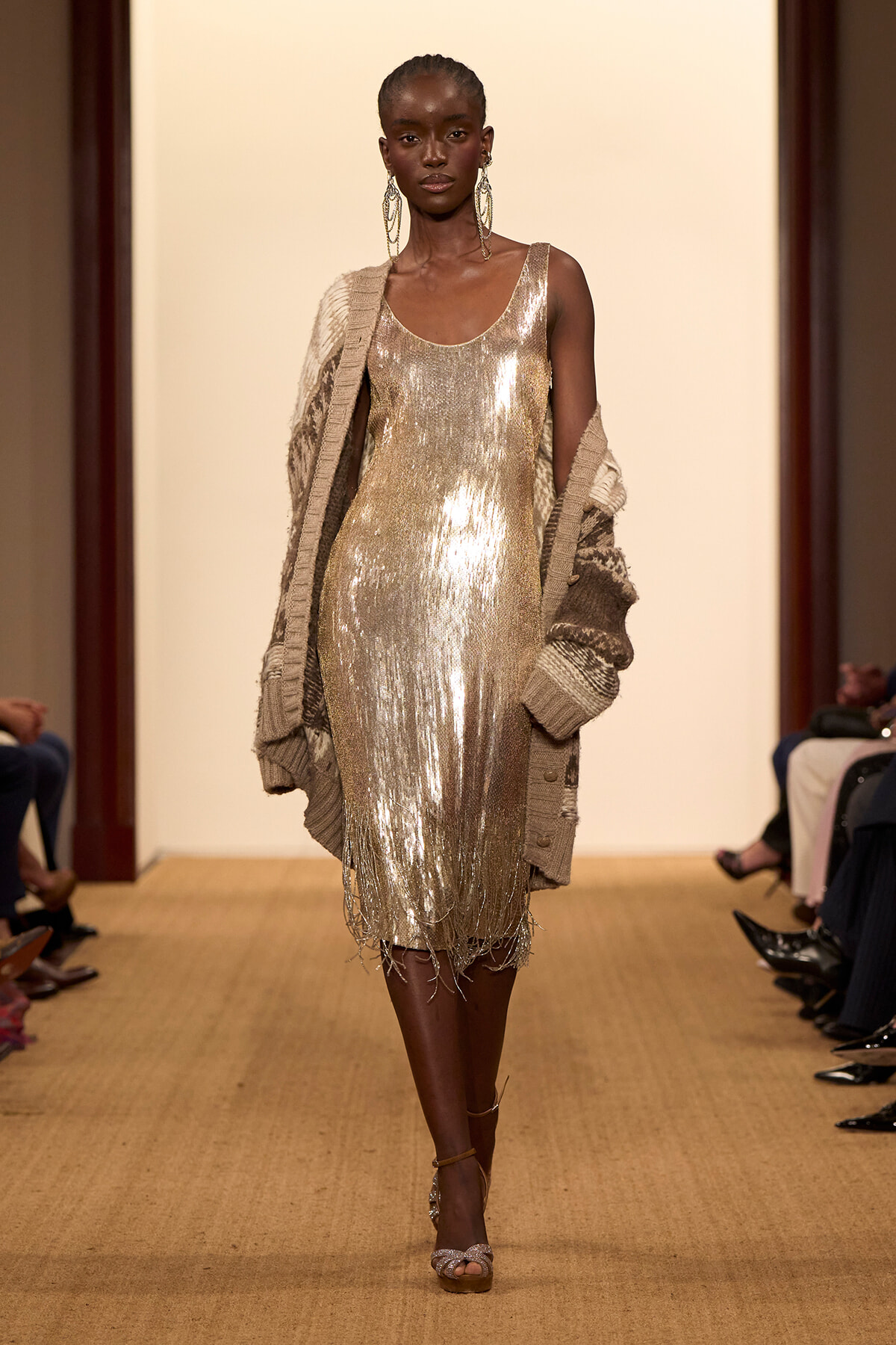 Model walking runway in a shiny gold sequin fringe dress with a chunky knit cardigan draped over shoulders.