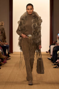 Model walking a runway in a brown turtleneck, fringed fur vest, and suede pants with a large belt buckle; audience seated along the aisle on both sides.
