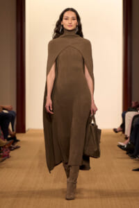 Model walking a runway in a brown ribbed turtleneck cape dress with a matching brown handbag and taupe boots, geometric earrings visible.