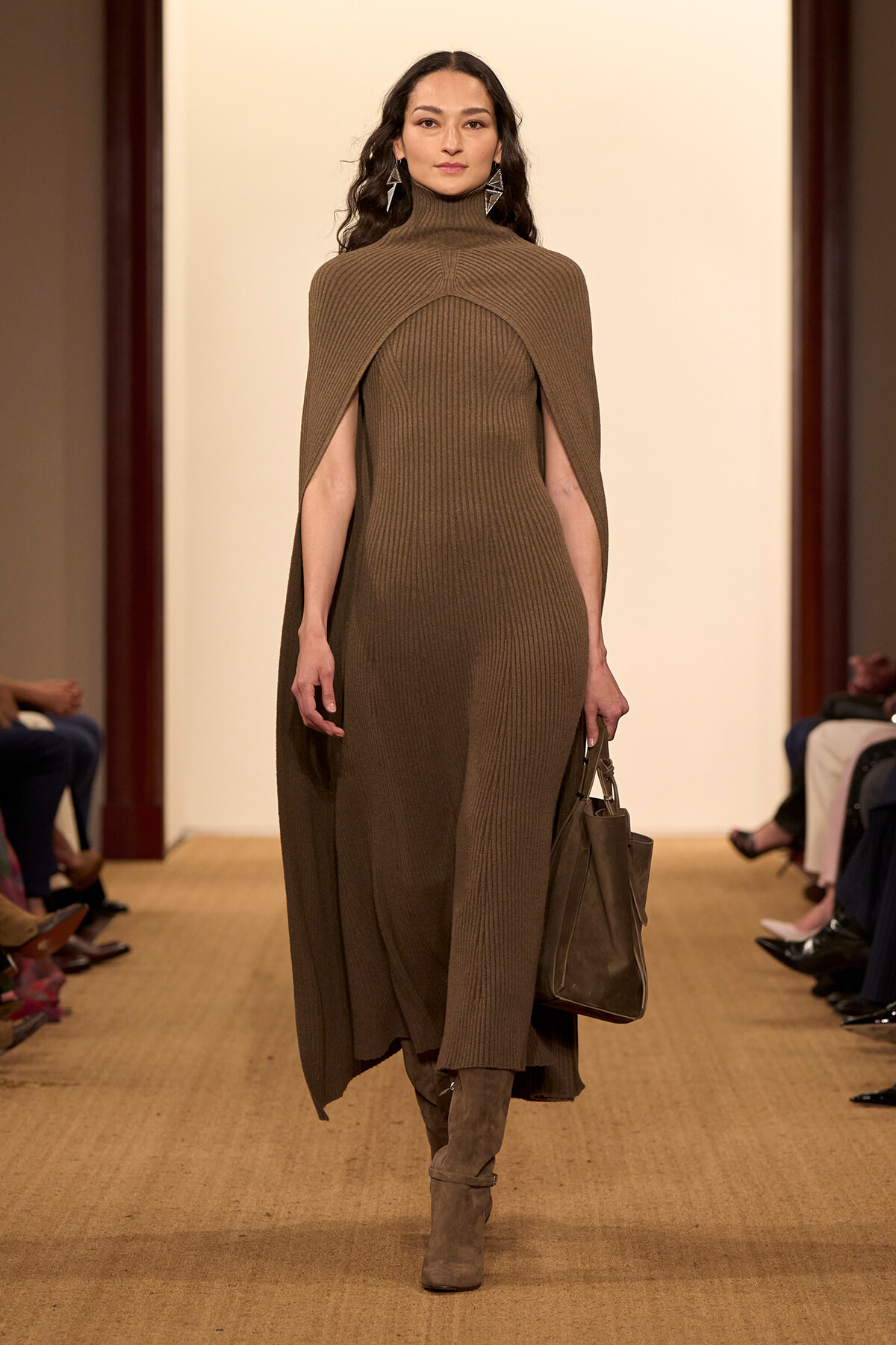 Model walking a runway in a brown ribbed turtleneck cape dress with a matching brown handbag and taupe boots, geometric earrings visible.