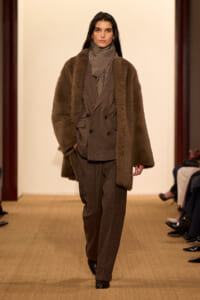 Model walking runway in a brown plaid suit with a matching scarf and a long brown faux-fur coat.