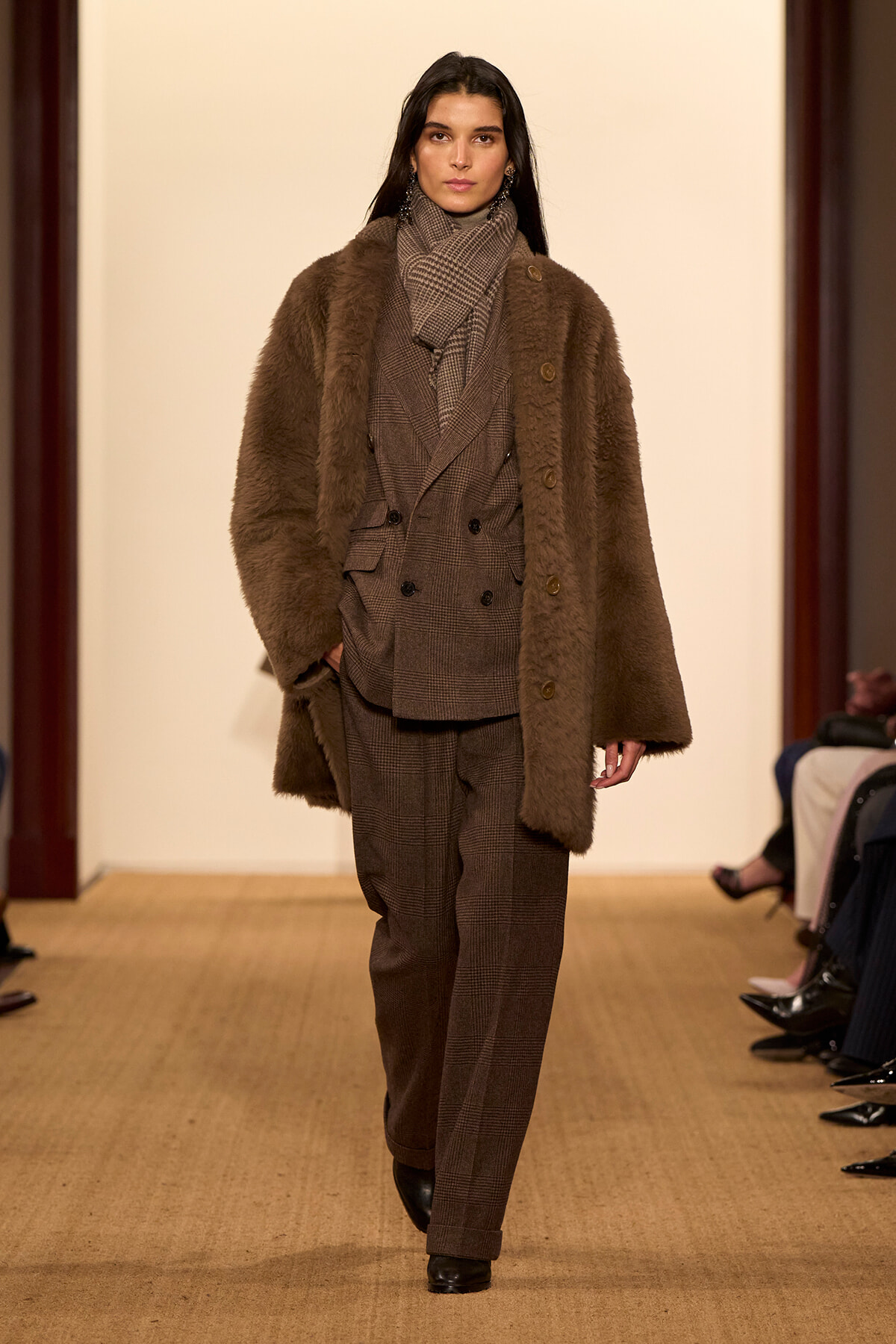 Model walking runway in a brown plaid suit with a matching scarf and a long brown faux-fur coat.