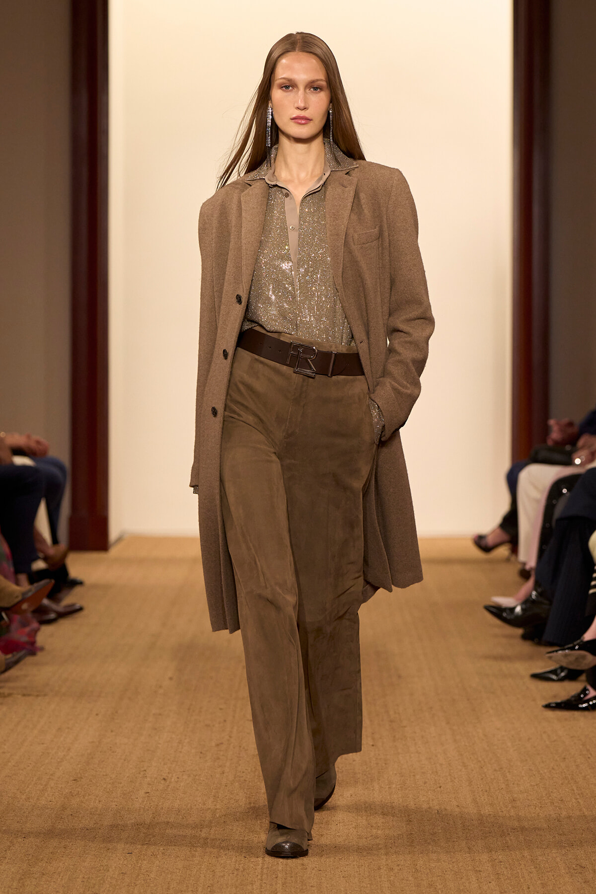 Model walking a fashion show runway in a tan coat over a sequined beige blouse and wide-leg brown pants with a dark brown belt and statement earrings.