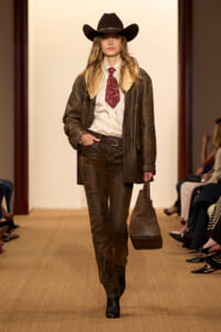 Female model walking the runway in a western-inspired outfit: black cowboy hat, shearling jacket, white shirt, patterned red tie, brown leather pants, and boots, carrying a brown bag.
