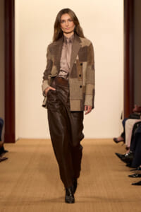 Model on a runway wearing a patchwork brown blazer, taupe satin shirt, tie, and dark brown leather pants.