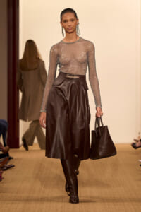 Model walks a fashion runway in a sparkly sheer long-sleeve top tucked into a high-waisted dark brown leather skirt, carrying a matching brown tote bag.