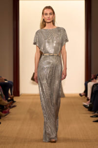 Model walking runway in a full-length silver sequined jumpsuit with a thin belt, neutral backdrop behind her.