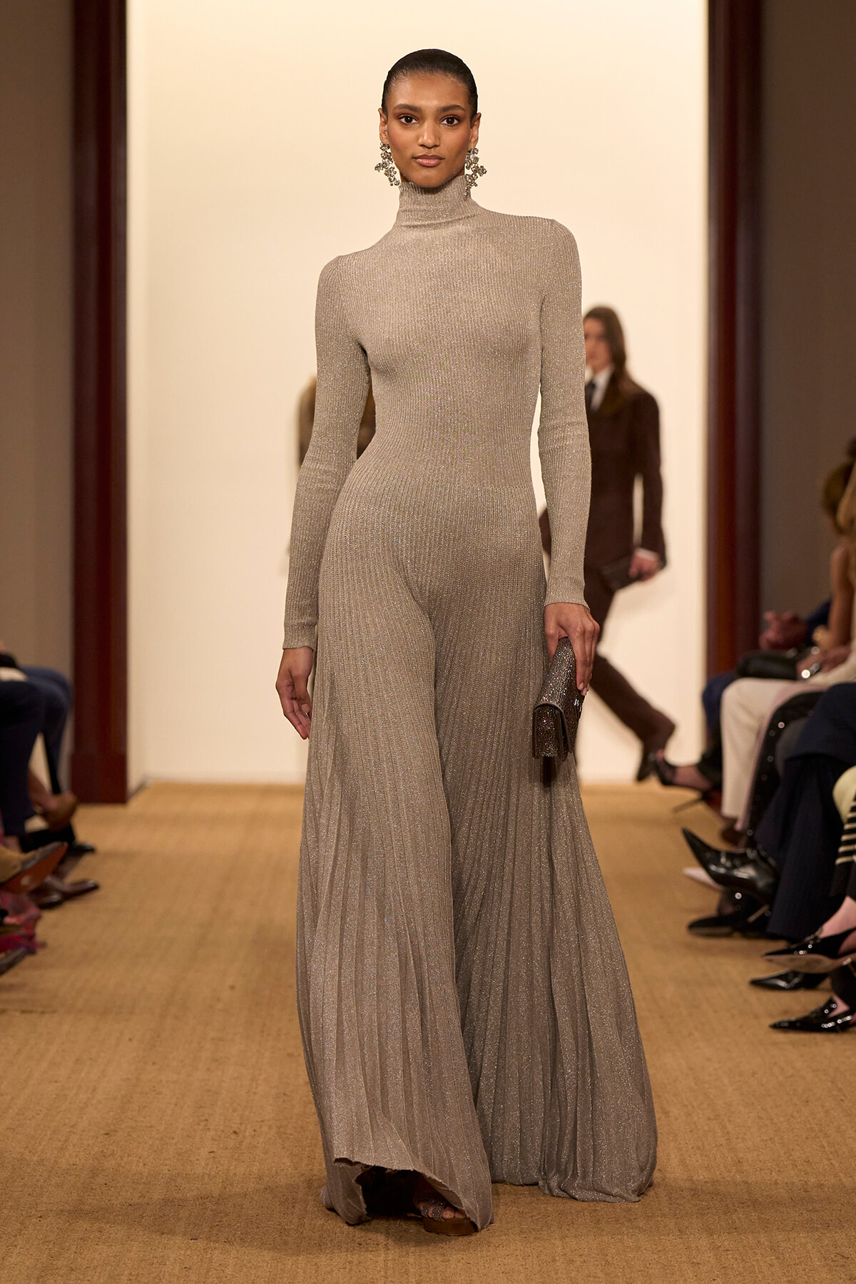 Model walks a runway in a shimmering beige turtleneck jumpsuit with wide legs, wearing statement chandelier earrings and a glitter clutch, audience in background.