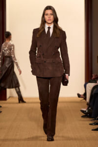Model walking a runway wearing a dark brown velvet double-breasted suit with white shirt and black tie, holding a small clutch.