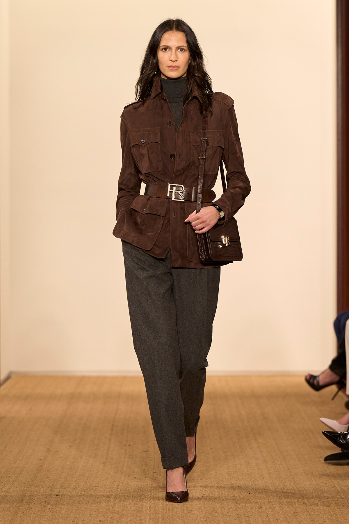 Model walking a runway in a brown suede utility jacket with a cinched waist, dark turtleneck, gray trousers, and a small black shoulder bag.