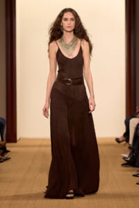 Model walking a runway in a dark brown sleeveless maxi dress with a wide belt and layered statement necklace, on a beige-lit catwalk.