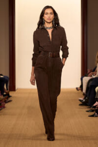 Model walking a fashion runway in a brown suede jumpsuit with a wide belt and silver choker, hands in pockets, neutral makeup.