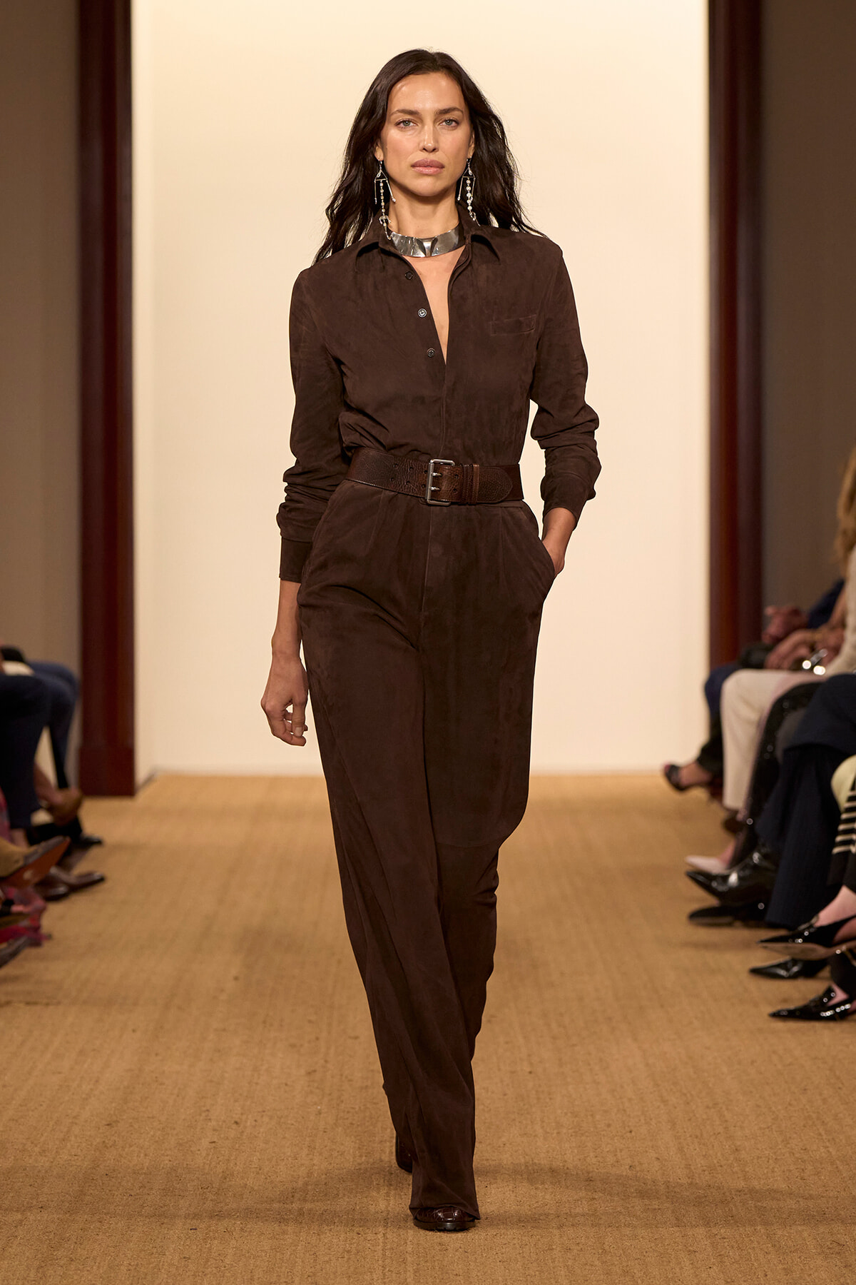 Model walking a fashion runway in a brown suede jumpsuit with a wide belt and silver choker, hands in pockets, neutral makeup.