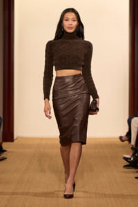Model walking a runway in a brown cropped turtleneck sweater and a high-waisted chocolate leather pencil skirt, holding a small clutch.