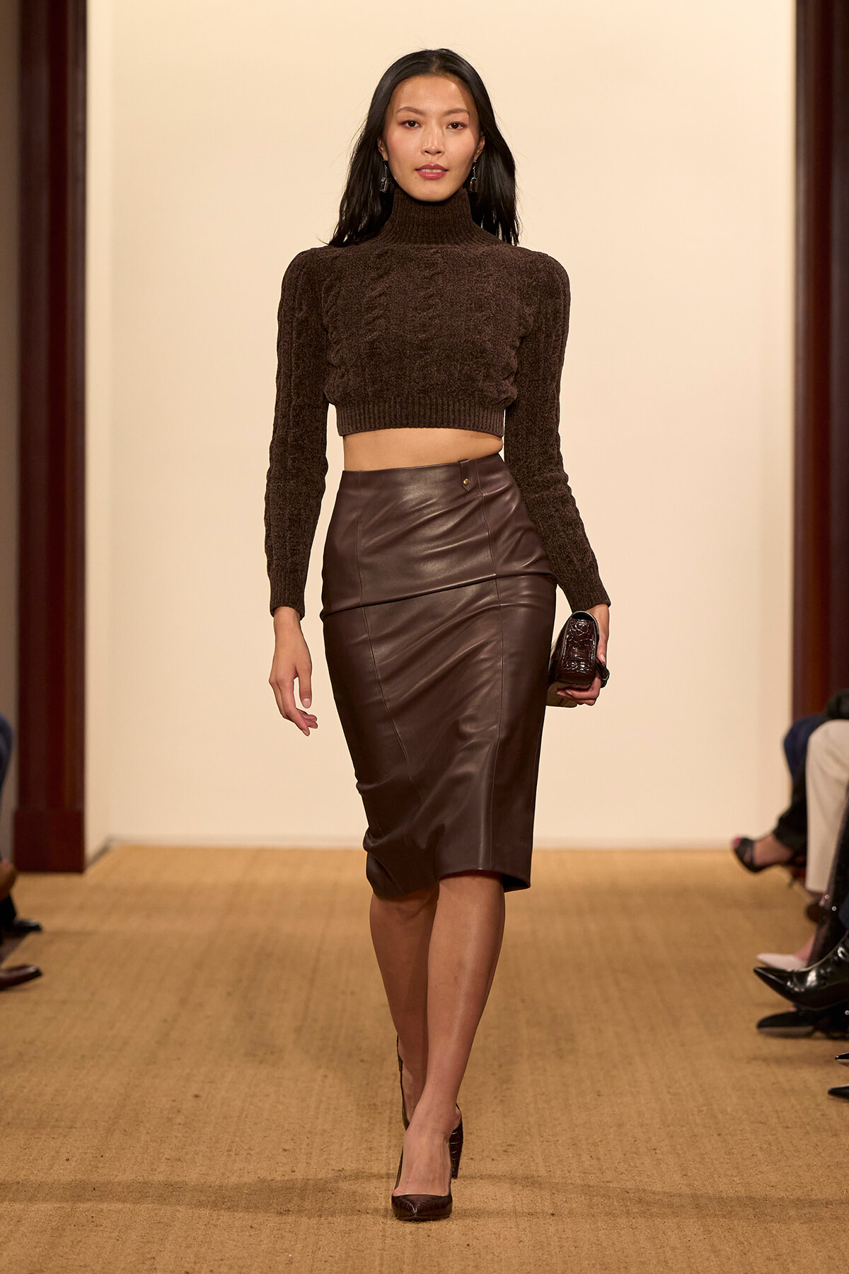 Model walking a runway in a brown cropped turtleneck sweater and a high-waisted chocolate leather pencil skirt, holding a small clutch.