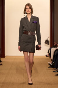Model walking a fashion runway in a dark gray blazer dress with a wide brown belt and purple pocket square, holding a small clutch.