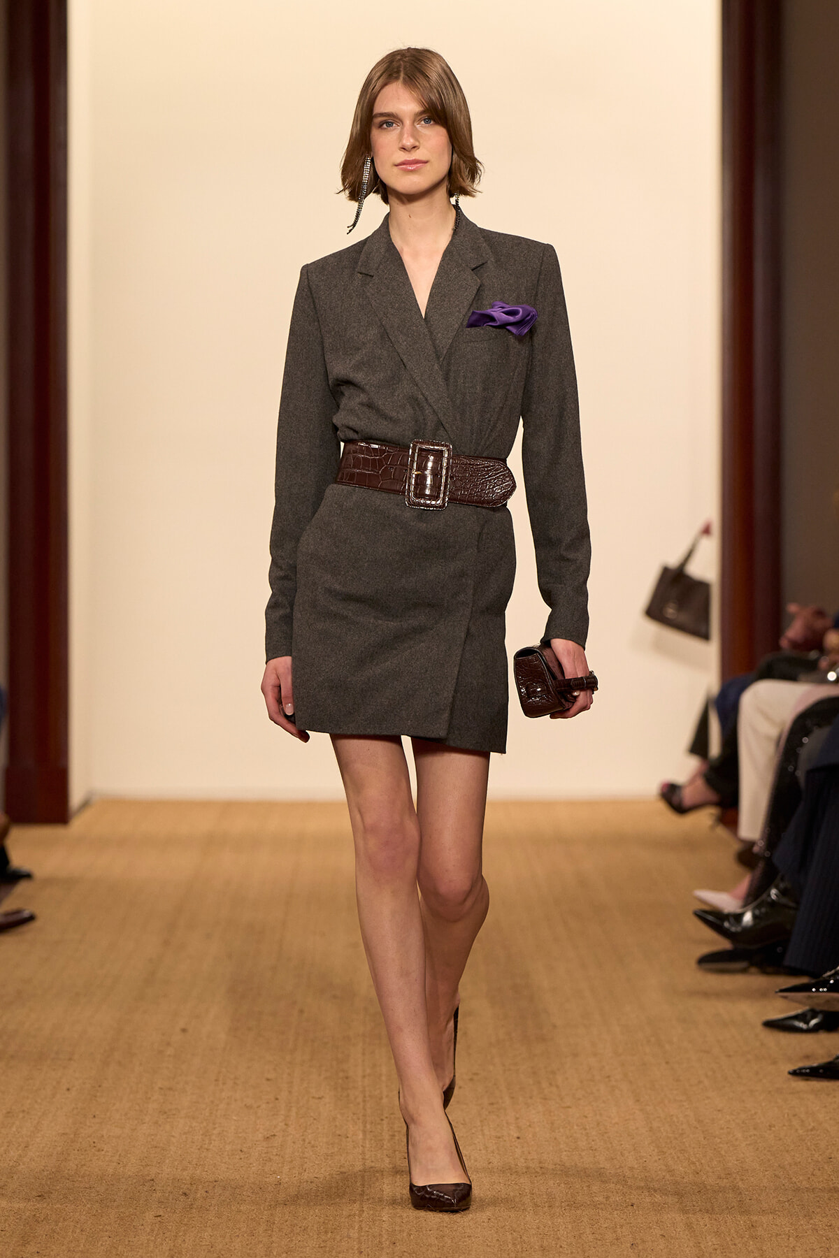 Model walking a fashion runway in a dark gray blazer dress with a wide brown belt and purple pocket square, holding a small clutch.