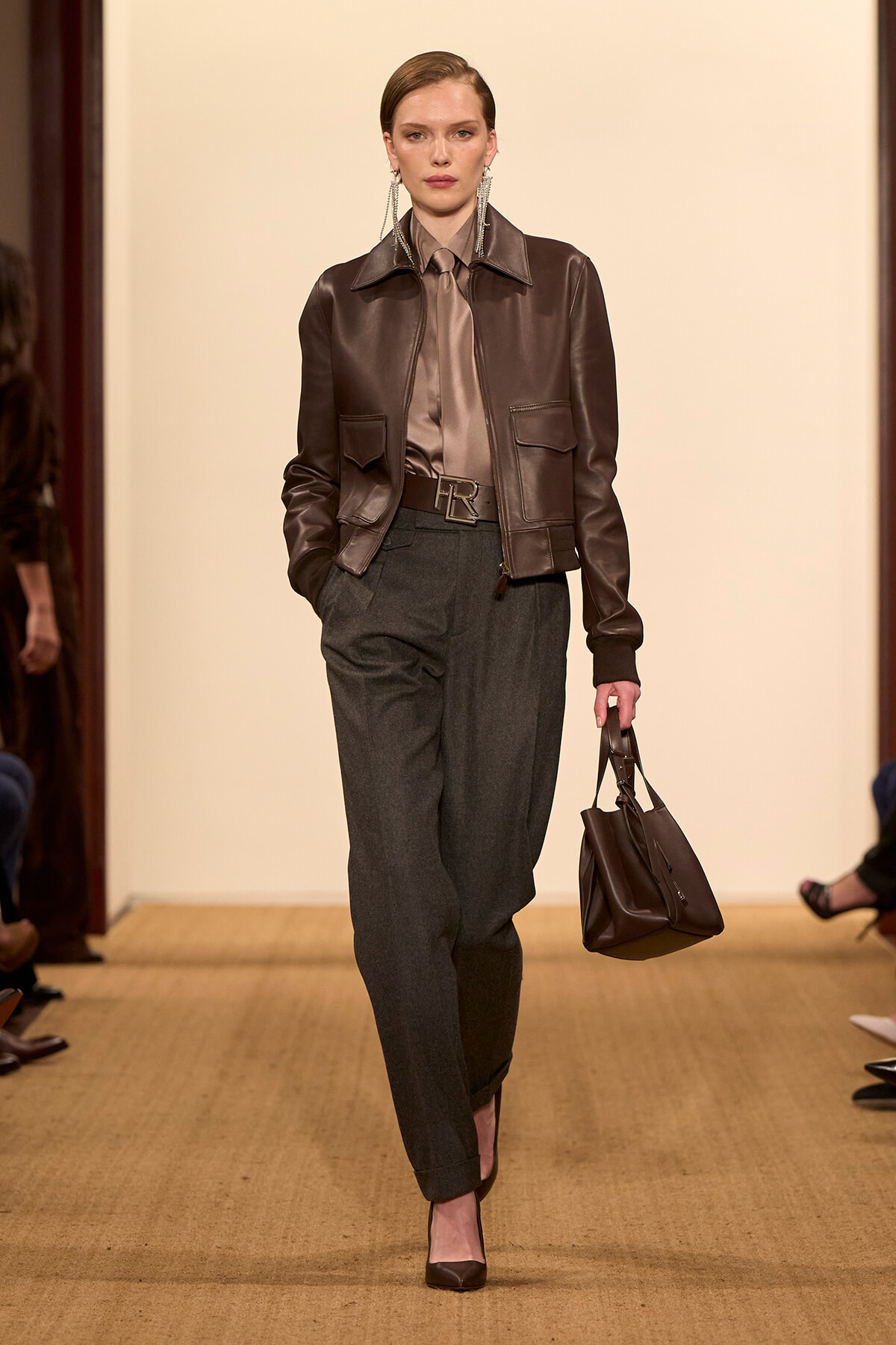 Model walking a runway in a brown leather jacket over a taupe shirt and matching tie, with gray tailored trousers and a brown handbag.