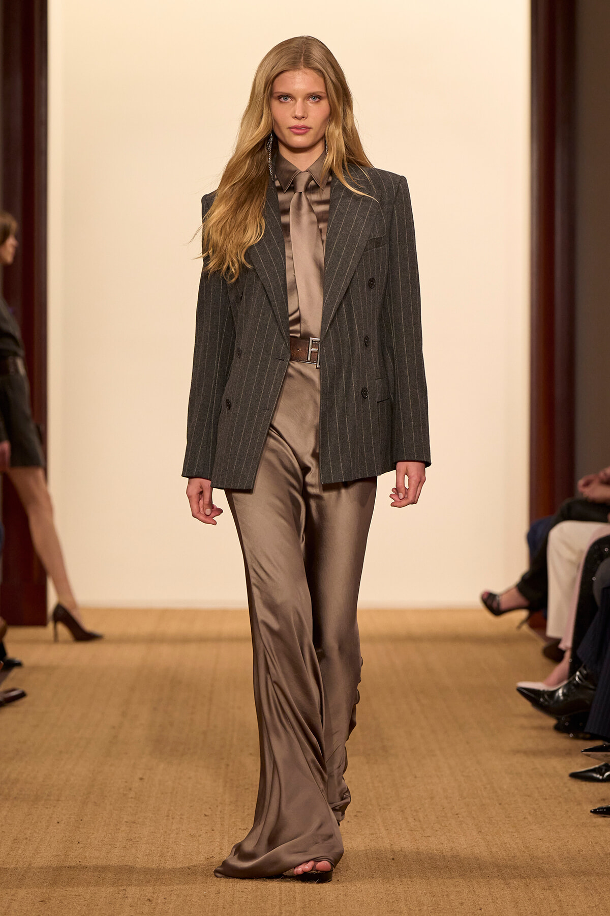 Model walking the runway in a charcoal pinstripe blazer over a taupe satin shirt and matching wide‑leg satin pants with a brown belt.