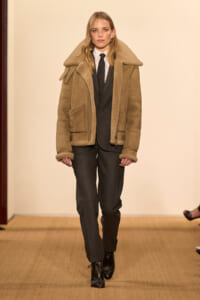 Female model walking a runway in a tan shearling jacket over a black suit with a white shirt and black tie.