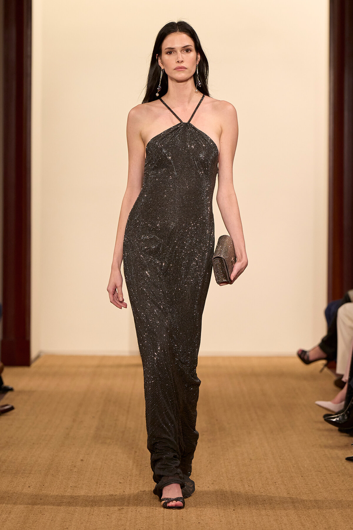 Model walks a runway in a shimmering black halter gown, holding a glittery clutch and wearing dangling earrings.