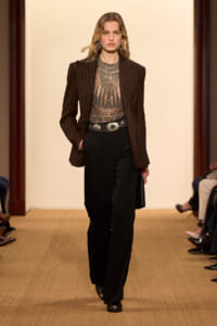 Model walking a runway in a dark pinstriped blazer, sheer beaded top, wide belt, and black trousers.