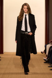Model walking runway in a black coat over a white shirt and black tie, paired with black fringe-detail pants and a silver belt buckle.